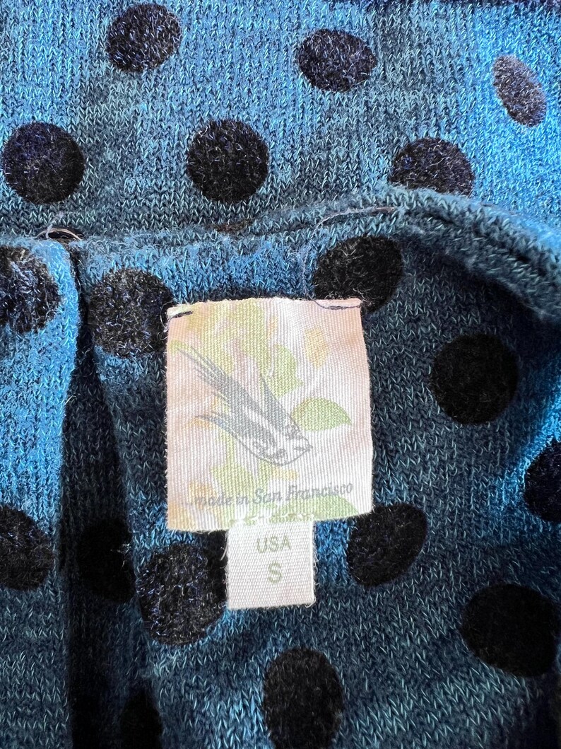 May include: Close-up of a blue knit fabric with large black polka dots. A tag is visible, with a bird illustration and text that reads "Made in San Francisco" and "USA S". The fabric appears to be a top or dress.
