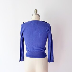 Op de afbeelding: Een periwinkelblauwe cardigan met lange mouwen, gepresenteerd op een paspop. De trui heeft een getailleerd silhouet en delicate details. De halslijn heeft subtiele ruches, wat een vleugje elegantie toevoegt. De paspop is van standaard formaat.