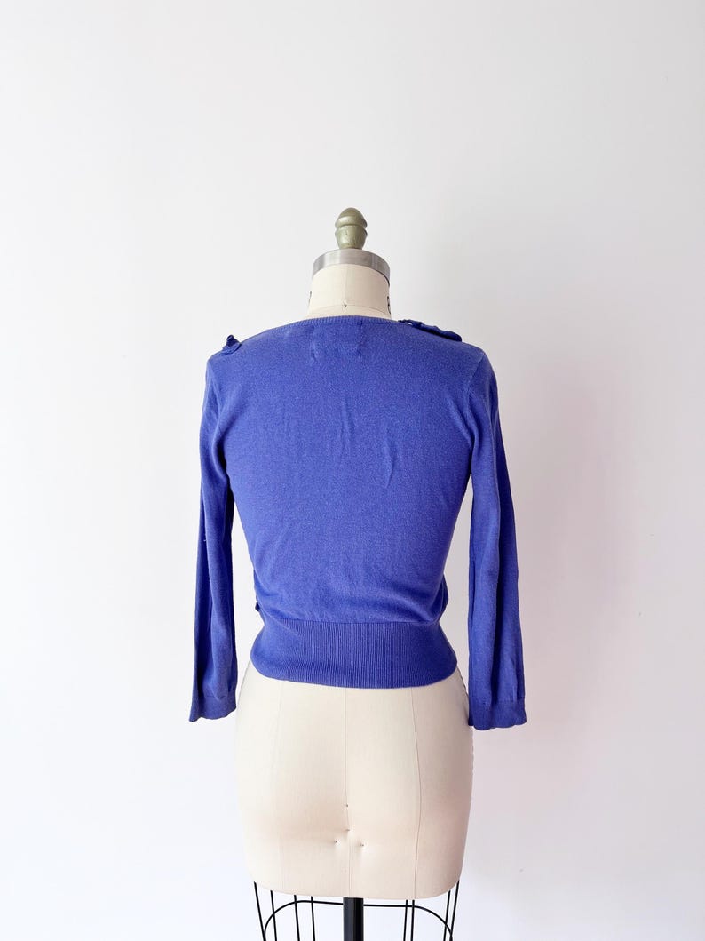 Op de afbeelding: Een periwinkelblauwe cardigan met lange mouwen, gepresenteerd op een paspop. De trui heeft een getailleerd silhouet en delicate details. De halslijn heeft subtiele ruches, wat een vleugje elegantie toevoegt. De paspop is van standaard formaat.