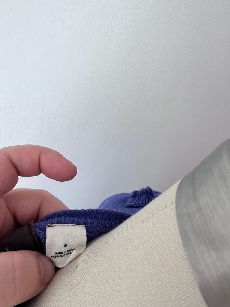 Op de afbeelding: Een close-up van een paars kledingstuk met een label met de tekst "S" en "MADE IN CHINA FABRIQUE EN CHINE". Het kledingstuk staat op een mannequin. Het label wordt door een hand vastgehouden.