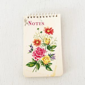 May include: A spiral-bound notepad with a cream-colored cover featuring a floral illustration of red, pink, and yellow roses and other blossoms. The word Notes is printed in red text at the top left. The notepad has a white plastic coil binding.