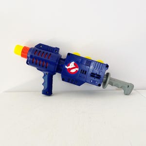 Op de afbeelding: Een blauw en grijs speelgoed proton pack met een gele spuitmond en handvat. Het speelgoed heeft het Ghostbusters-logo en rode accenten. Het speelgoed is gemaakt van plastic en is ontworpen om op een wapen te lijken.