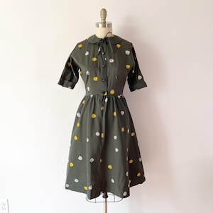 Op de afbeelding: Een olijfgroene vintage jurk met een kraag en korte mouwen. De jurk heeft een knoopsluiting aan de voorkant en is versierd met gele en witte abstracte vormen. De jurk wordt op een mannequin gepresenteerd.