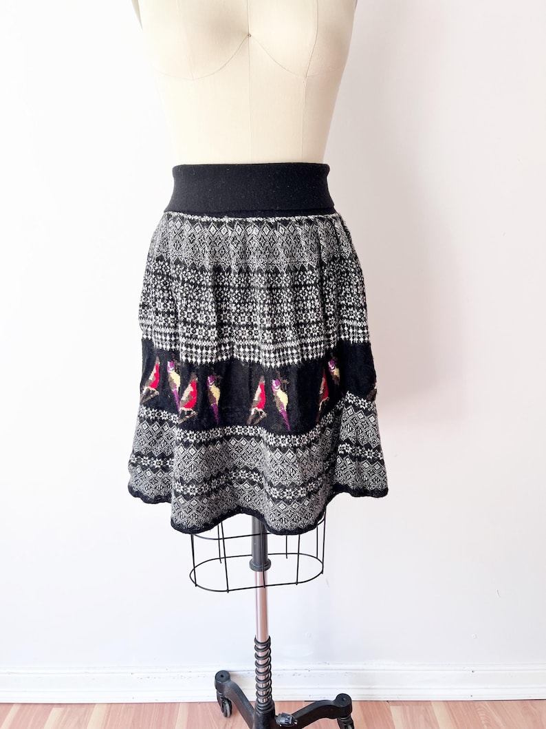 May include: A black and white patterned skirt with a black waistband and hem. The skirt features a repeating geometric design and a row of colorful bird motifs. The skirt is displayed on a mannequin.