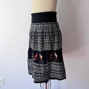 May include: A black and white patterned skirt with a wide black waistband. The skirt features a detailed knit design with a band of colorful bird motifs. The skirt is displayed on a mannequin.