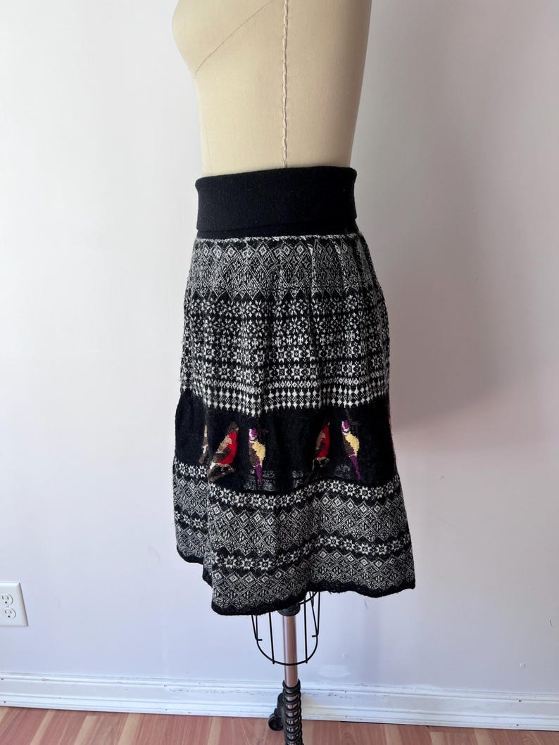 May include: A black and white patterned skirt with a wide black waistband. The skirt features a detailed knit design with a band of colorful bird motifs. The skirt is displayed on a mannequin.