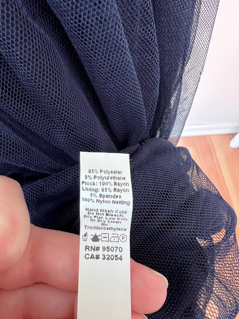 May include: Close-up of navy blue netting fabric. A white tag lists the fabric composition as 95% polyester, 5% polyurethane, 100% rayon flock, 95% rayon lining, 5% spandex, and 100% nylon netting. Care instructions are also listed.