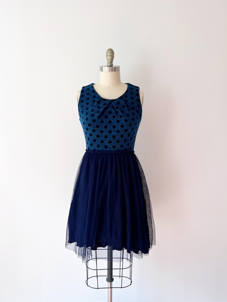 May include: A sleeveless blue dress with black polka dots on the bodice and a navy blue tulle skirt. The dress is displayed on a mannequin. The bodice has a small collar.