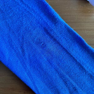 Op de afbeelding: Close-up van een levendige blauwe stof, mogelijk een mouw of een deel van een kledingstuk. Het materiaal heeft een getextureerd uiterlijk met subtiele kleurvariaties en zichtbare vezels. De stof is effen, verzadigd blauw.