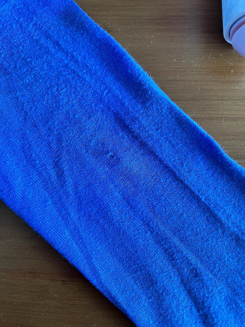 Op de afbeelding: Close-up van een levendige blauwe stof, mogelijk een mouw of een deel van een kledingstuk. Het materiaal heeft een getextureerd uiterlijk met subtiele kleurvariaties en zichtbare vezels. De stof is effen, verzadigd blauw.