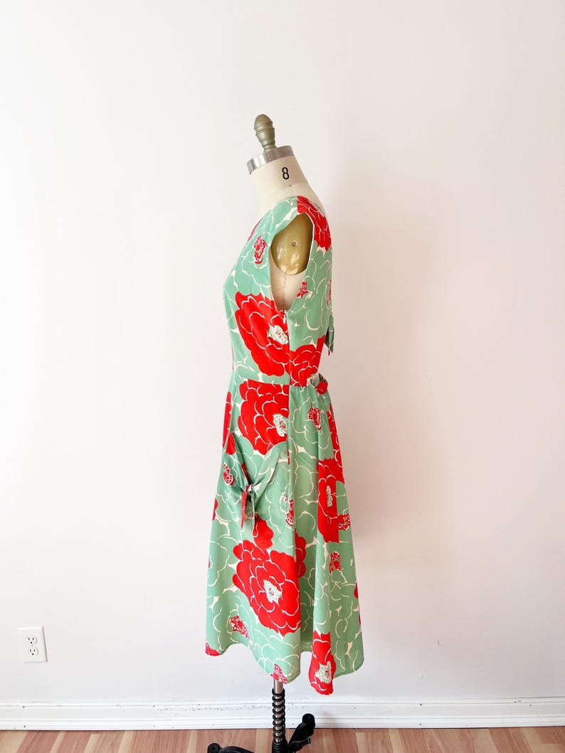 Może przedstawiać: Sukienka w stylu vintage na manekinie. Sukienka ma jasne zielone tło z dużymi czerwonymi wzorami kwiatowymi. Ma dekolt w serek, kr&oacute;tkie rękawy i sp&oacute;dnicę do kolan. Sukienka ma kieszenie z detalami w postaci kokardek.