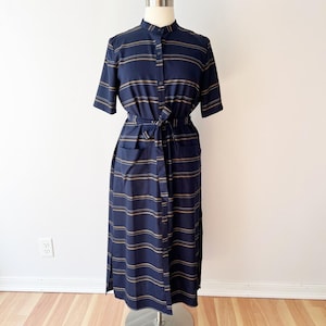 May include: A navy blue midi dress with horizontal tan stripes. The dress has short sleeves, a button-up front, a mandarin collar, and a matching tie belt. It features two front pockets and is displayed on a mannequin.