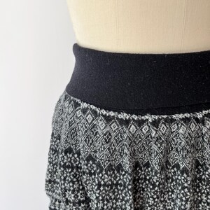 May include: A black and white patterned skirt with a wide black waistband. The skirt features a geometric design in shades of gray and black, with a hint of color at the bottom. The skirt is displayed on a mannequin.