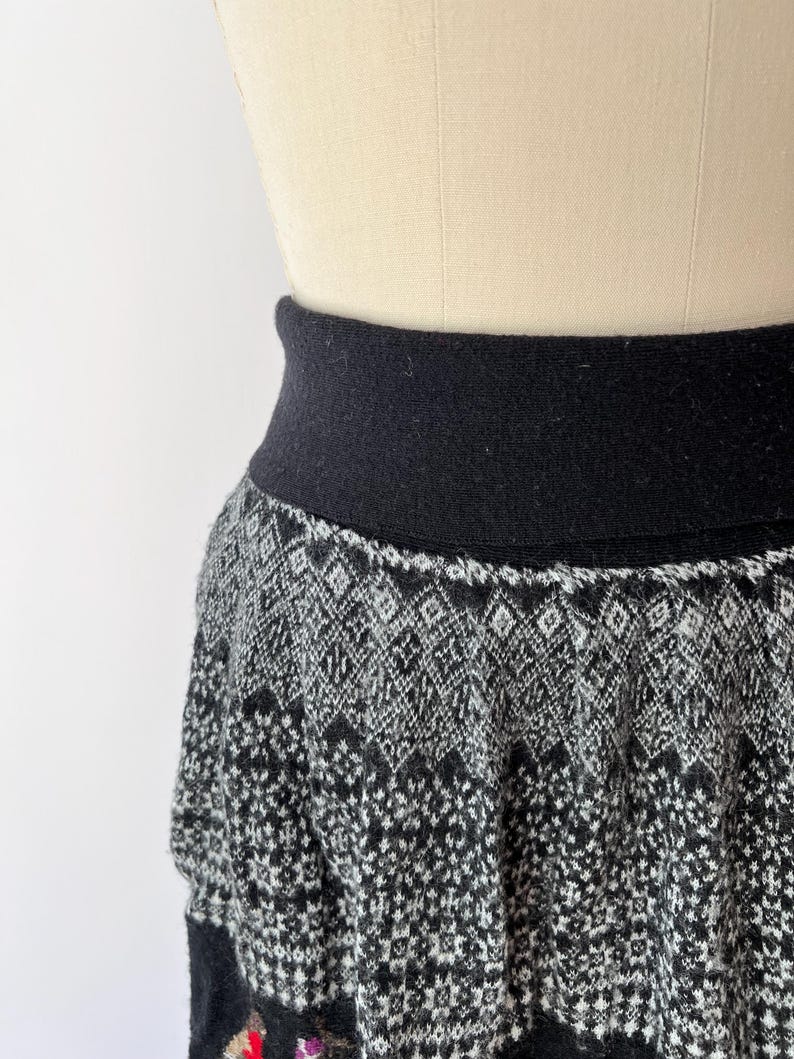 May include: A black and white patterned skirt with a wide black waistband. The skirt features a geometric design in shades of gray and black, with a hint of color at the bottom. The skirt is displayed on a mannequin.
