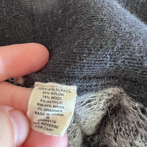 May include: Close-up of a tag with fabric composition details: 41% Alpaca, 35% Nylon, 19% Wool, 5% Acrylic, and 5% Spandex. The tag also indicates the item is made in China. The garment is a dark gray color.