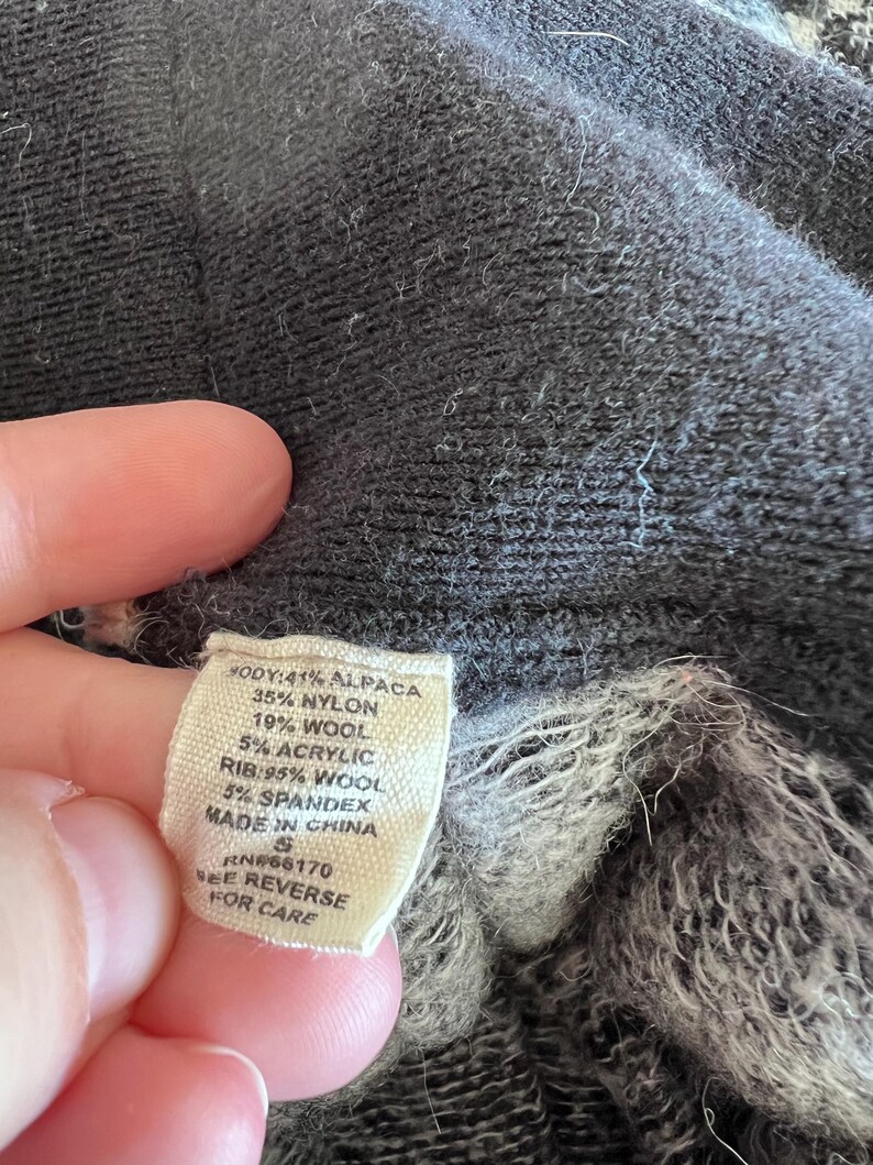 May include: Close-up of a tag with fabric composition details: 41% Alpaca, 35% Nylon, 19% Wool, 5% Acrylic, and 5% Spandex. The tag also indicates the item is made in China. The garment is a dark gray color.