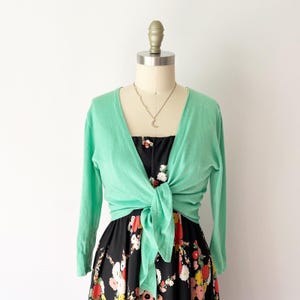 May include: A light green long-sleeve cardigan with a front tie closure, worn over a black dress featuring a colorful floral pattern of white, red, and yellow flowers. A delicate silver-toned necklace with a crescent moon pendant is visible.