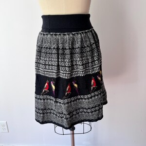 May include: A black and white patterned skirt with a wide black waistband. The skirt features a repeating geometric design and a band of colorful bird motifs. The skirt is displayed on a mannequin.