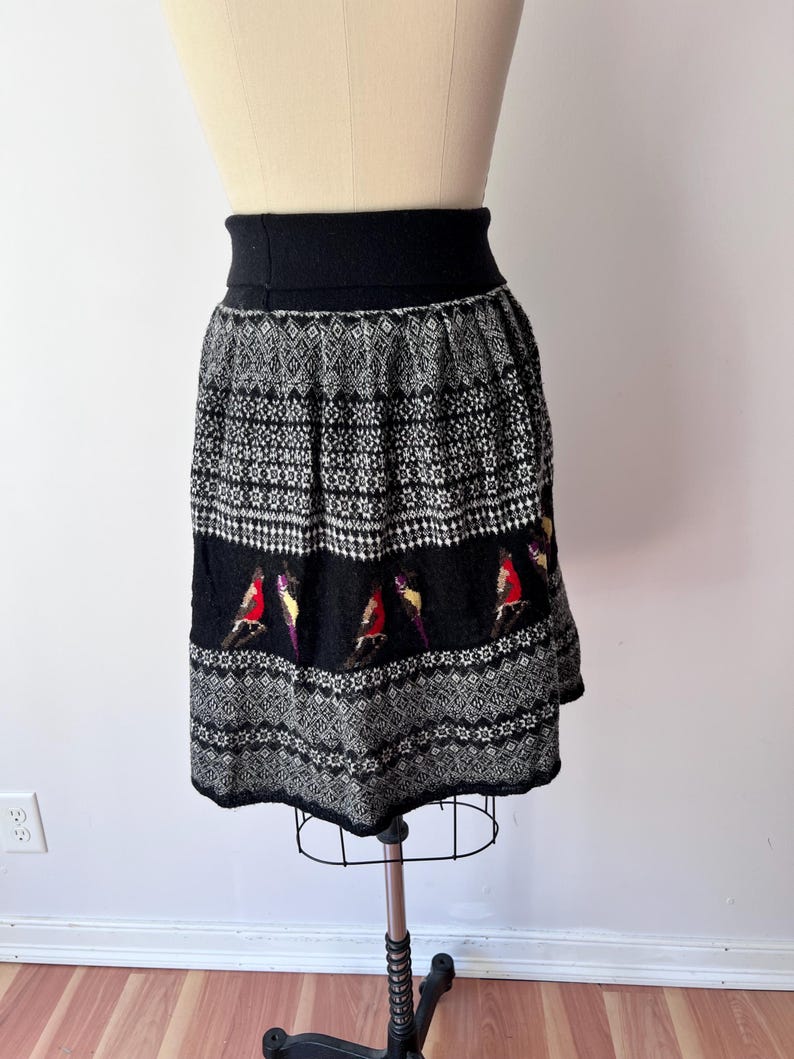 May include: A black and white patterned skirt with a wide black waistband. The skirt features a repeating geometric design and a band of colorful bird motifs. The skirt is displayed on a mannequin.