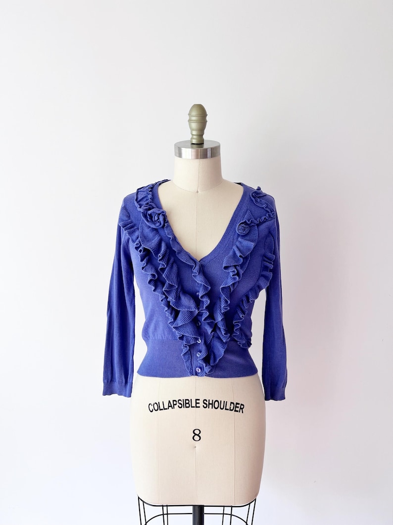Op de afbeelding: Een periwinkle blauw vest met een V-hals en lange mouwen. De voorkant heeft een trapsgewijs ruchesontwerp. Het kledingstuk wordt getoond op een paspop met de tekst "COLLAPSIBLE SHOULDER" en het nummer "8".