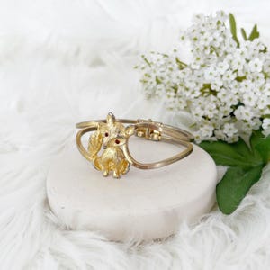 May include: Gold-toned fox bracelet with red rhinestone eyes. The hinged bracelet features a detailed fox charm with a textured finish. The bracelet is displayed on a white surface with a soft, white background and white flowers.