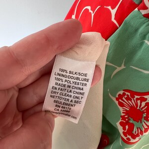 Może przedstawiać: Zbliżenie na metkę odzieżową z tekstem. Na metce widnieje napis "100% JEDWAB/SOIE PODSZEWKA/DOUBLURE 100% POLIESTER MADE IN CHINA EN FAIT LA CHINE CZYŚCIĆ CHEMICZNIE NETTOYER A SEC SEULEMENT RN 66170 CA 32064." Odzież ma czerwony i zielony wz&oacute;r kwiatowy.
