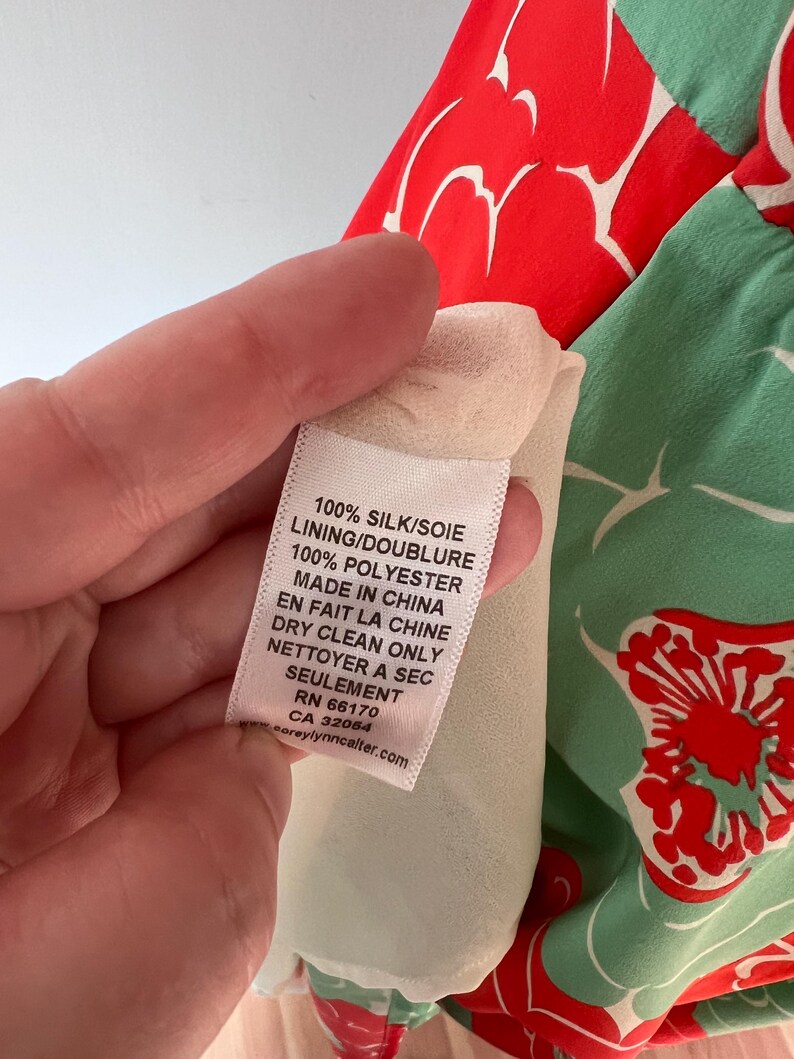 Może przedstawiać: Zbliżenie na metkę odzieżową z tekstem. Na metce widnieje napis "100% JEDWAB/SOIE PODSZEWKA/DOUBLURE 100% POLIESTER MADE IN CHINA EN FAIT LA CHINE CZYŚCIĆ CHEMICZNIE NETTOYER A SEC SEULEMENT RN 66170 CA 32064." Odzież ma czerwony i zielony wz&oacute;r kwiatowy.