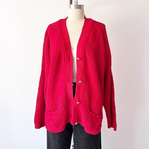 May include: A bright pink cardigan sweater with a ribbed knit texture. The sweater has a V-neck, long sleeves, two front pockets, and three silver-toned buttons. The garment is displayed on a mannequin.