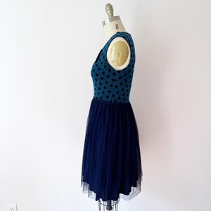 May include: A teal dress with black polka dots on the bodice and a navy blue tulle skirt. The dress is displayed on a dress form. The dress has a V-neck and a fitted waist. The tulle skirt falls to the mid-calf.