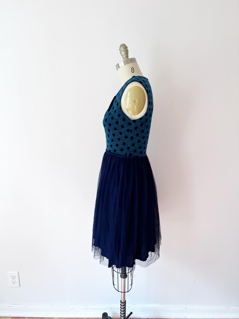 May include: A teal dress with black polka dots on the bodice and a navy blue tulle skirt. The dress is displayed on a dress form. The dress has a V-neck and a fitted waist. The tulle skirt falls to the mid-calf.