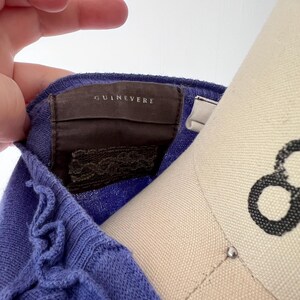 Op de afbeelding: Close-up van een periwinkle blauwe gebreide top met een gerimpelde rand. Het label leest "GUINEVERE" in een schreefletter. De top is over een mannequin gedrapeerd en toont de textuur en kleur van de stof.