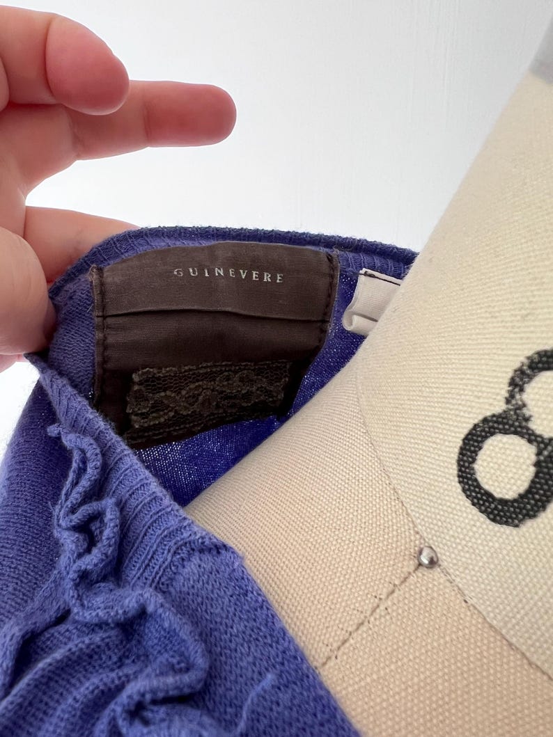 Op de afbeelding: Close-up van een periwinkle blauwe gebreide top met een gerimpelde rand. Het label leest "GUINEVERE" in een schreefletter. De top is over een mannequin gedrapeerd en toont de textuur en kleur van de stof.