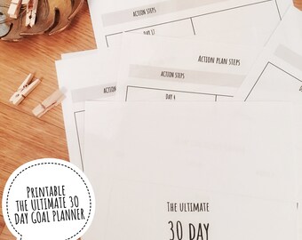 Könnte beinhalten: Ein Stapel weißer Papier-Zielplaner mit dem Text "The Ultimate 30 Day Goal Planner" und "Action Plan Steps". Das Bild enthält eine goldfarbene Blattschale und mehrere kleine Wäscheklammern aus Holz.
