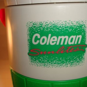 May include: White Coleman Sunlite cooler with a green and white logo. The cooler has red handles and a green base.