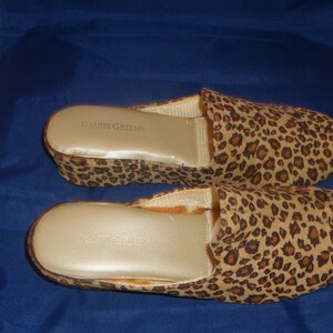 May include: A pair of leopard print house slippers with a wedge heel. The slippers are made by the brand "Camille Green".