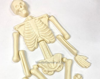 May include: A white chocolate skeleton made of individual pieces, including the skull, rib cage, pelvis, arms, legs, hands, and feet.  The pieces are laid out on a white surface.