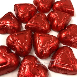 Peut inclure: Un assortiment de chocolats en forme de cœur, enveloppés dans du papier d'aluminium rouge brillant. Les bonbons sont disposés sur une surface blanche, leurs emballages brillants reflétant la lumière. L'image est un gros plan, mettant en valeur la texture et la couleur du papier.