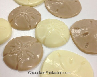 May include: An assortment of sand dollar-shaped chocolates in shades of ivory and light brown. The chocolates have detailed impressions and a glossy finish. The text "ChocolateFantasties.com" is visible in the image.