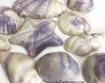 May include: A collection of white chocolate seashells and a starfish, all with purple marbling. The chocolates are arranged on a white surface. ChocolateFantasies.com IslandGirlGourmet on Etsy