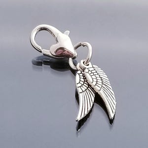 May include: A silver-colored charm featuring a lobster clasp and two small wing pendants. The wings are detailed with feather patterns. The charm is designed for jewelry making or as a decorative accessory.