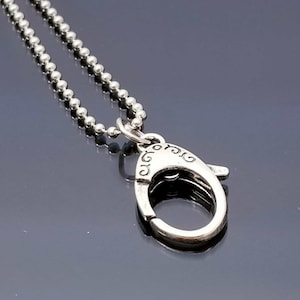 May include: A silver-colored necklace with a ball chain and a decorative clasp. The clasp has an oval shape with etched designs. The necklace is displayed on a reflective surface.