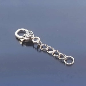 May include: A silver-toned heart-shaped clasp with an ornate design, connected to a chain of interlocking links and a small ring. The clasp is likely for jewelry or accessories.