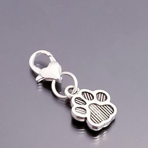 May include: A silver-colored charm featuring a paw print design. The charm is attached to a clasp, suitable for attaching to a keychain, necklace, or bracelet. The paw print has detailed lines.