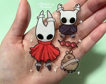 May include: A clear sticker sheet featuring four cartoon character designs. The characters include a figure in a red cape, a figure in a purple cloak, a small brown creature, and a string of red beads. The sticker sheet has the text "Sinnin Studios".