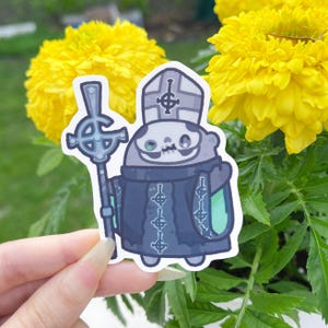 May include: A sticker featuring a cartoon character in a dark blue robe and a skull-like face, holding a silver staff with a cross. The character wears a tall, white hat. The background includes bright yellow flowers and green foliage.