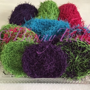 May include: A collection of colorful, round, textured scrubbies in shades of purple, green, blue, and pink. The scrubbies are arranged on a clear, rectangular, glass dish. The image has a soft focus.