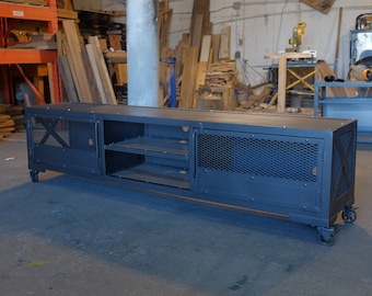 May include: A black metal entertainment center with two doors, two shelves, and a wire mesh design. The entertainment center is on wheels and has a rustic industrial style.