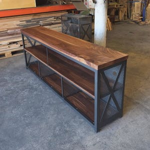 May include: A long, wooden shelf unit with a black metal frame and wire mesh shelves. The shelf is made of dark wood with a natural grain pattern. The metal frame has a black finish and is accented with X-shaped crossbars.