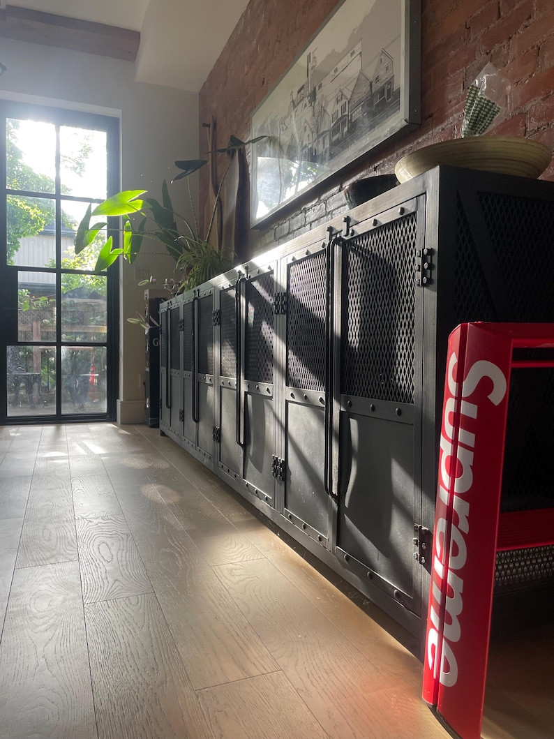 May include: A long, black metal cabinet with mesh door panels and riveted details. The cabinet is in a room with a brick wall, a framed picture, and a red Supreme chair. The room has a wooden floor and a door with sunlight streaming through.