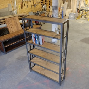 May include: A black metal bookshelf with five wooden shelves. The shelves are made of reclaimed wood and have a rustic look. The bookshelf is standing on a concrete floor in a workshop.